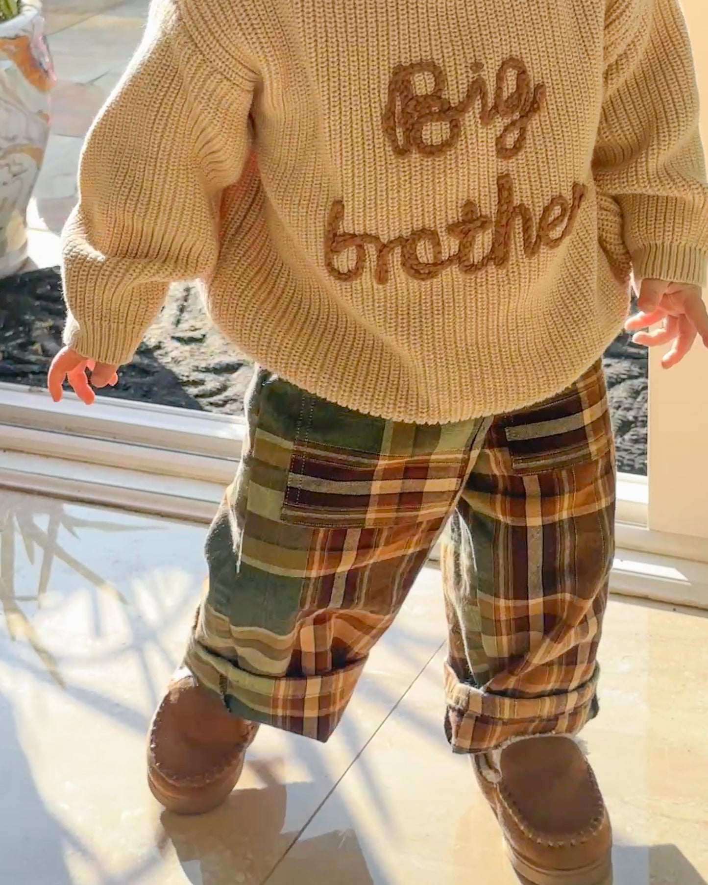 Toddler wearing the YANANAs toddler trousers with a beige knit sweater that says "big brother" and baby moccasins 